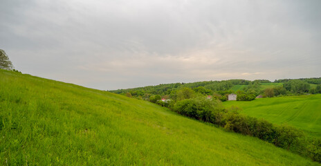 Mała wioska w dolinie wśród trawiastych wzgórz w Górach Świętokrzyskich. Pochmurne niebo nad piękną okolicą (koło Ostrowca) w wiosenne popołudnie. - obrazy, fototapety, plakaty