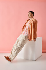Fashion shot of fashionable man in beige shirt, pants and boots on white cube on pink background