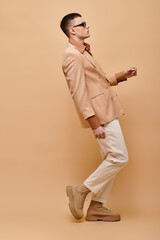 Side view photo of fancy man in beige jacket, shirt, pants and boots posing on beige background