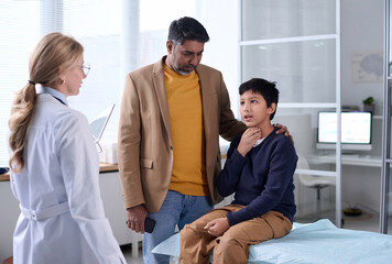 Portrait of Middle Eastern father and son visiting doctor in clinic with young boy explaining symptoms and pointing at throat