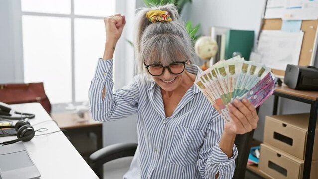 Triumphant middle age woman with grey hair ecstatically waves hong kong dollar banknotes in office victory celebration! raised arms displaying confident success, pure joy radiates.