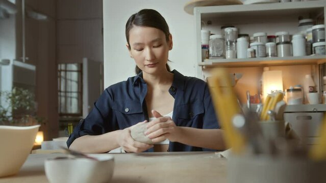 young Asian female artisan sits at table in ceramic studio. freelance ceramist shaping and forming clay for pottery.
