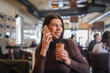 A one young happy girl or woman is drinking cold coffee in cafe or restaurant while using her phone...