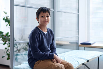 Portrait of young Middle Eastern boy sitting on cot in medical clinic and looking at camera with...