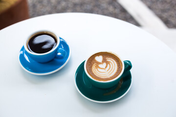 Two cups of coffee for breakfast on a white round table. Coffee for two. Cappuccino and black coffee in colored mugs - obrazy, fototapety, plakaty