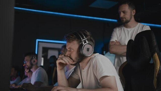 Cyber team coach is walking and watching game of his players in E-sport tournament. Gamers play video game on PC at computer club. 5 Caucasians of different ages in headset. Tactics strategy trainer