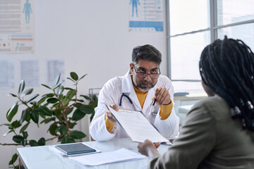 Portrait of senior Middle Eastern doctor handing documents to patient across table copy space