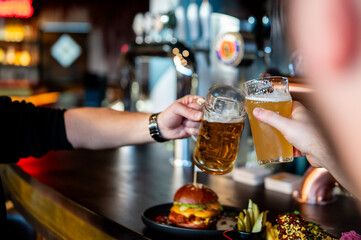 Two friends clinking beer glasses in a lively bar, with a burger and snacks in the foreground - obrazy, fototapety, plakaty