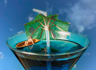 A miniature man sitting in a boat inside a cocktail glass.  A holiday or tourism concept.  A macro image. - obrazy, fototapety, plakaty