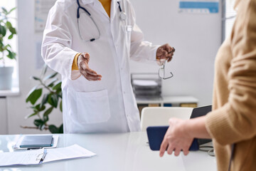 Close up of unrecognizable doctor greeting patient in medical clinic and gesturing for seat copy...