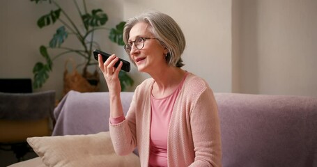Mature old 60s woman, older middle aged female customer holding smartphone using mobile app listening voice message and recording audio on cell phone technology device sitting on couch at home - Powered by Adobe