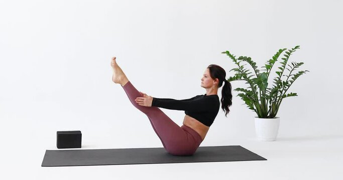 Athletic woman performs a variation of the Paripurna Navasana exercise, boat pose, practices yoga in leggings and a short long-sleeved T-shirt while sitting on a mat in a bright room