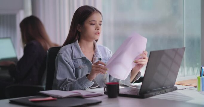 Indian Asian gen z young adult busy woman lady staff do pen paper work task sitting chair indoor job office use laptop teen girl intern female worker learn makes notes typing email check online data