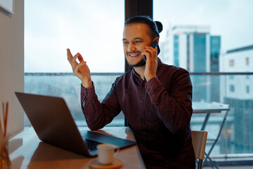 Portrait of smiling handsome man with hair bun working at laptop, talking on smartphone, on background of panoramic window.
