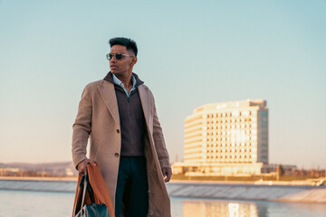 A man wearing a brown coat and sunglasses stands on a sidewalk next to a body of water. He is holding a brown bag and he is waiting for someone. The scene has a calm and relaxed mood