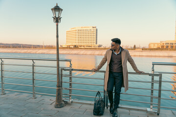 A man in a tan coat stands on a pier next to a body of water. He is holding a black bag and wearing a brown coat