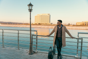 A man in a brown coat stands on a pier next to a body of water. He is holding a black bag and wearing glasses. The scene is peaceful and serene, with the man looking out over the water