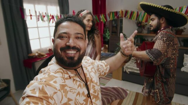 Medium UGC selfie shot of cheerful Mexican man holding out smartphone and filming himself and friends having fun at house party, guy in sombrero playing guitar, and girl dancing and swirling to music