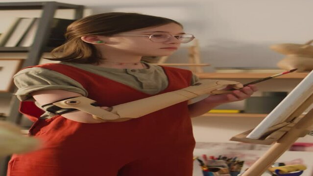 Vertical side footage of calm young girl in glasses and jumpsuit learning to paint with bionic arm holding brush and painting still life at home alone