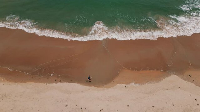 Drone footage of sea waves coming on a sandy beach with an adult man running in the early morning