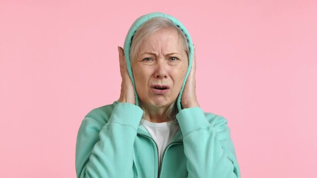 A distressed senior woman is covering her ears with her hands, showing a troubled reaction to a very loud and discomforting noise against a pale pink background. Camera 8K RAW. 