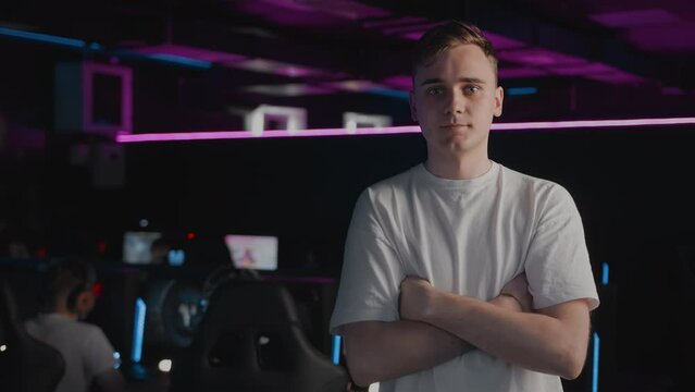 Portrait serious Caucasian youth gamer teenager poses, player crosses his arms and looks at camera in computer club, cyber sport tournament on PC. E-sport multiplayer online video games championship