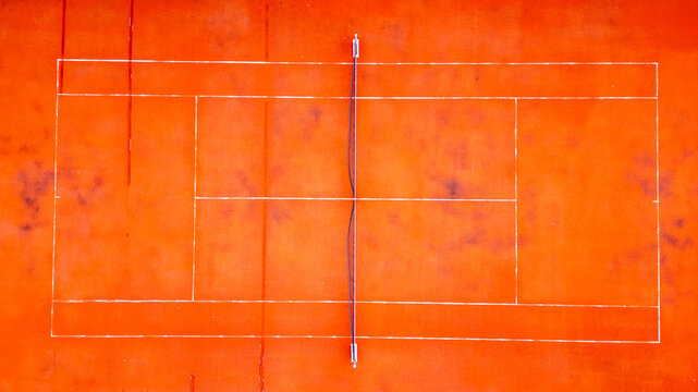 An aerial perspective showcases the geometric beauty of a vibrant orange tennis court, the bold white lines creating a stark contrast. The surface shows signs of weathering, adding texture and