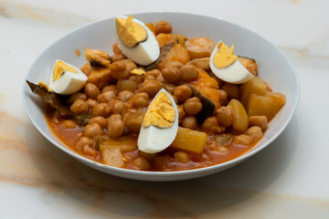 lent stew: a plate of chickpeas with pepper, potatoes and cod with tomato and hard-boiled egg on a white veined marble table