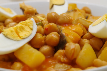 Lenten stew: detail of a plate of chickpeas with pepper, potatoes and cod with tomato and a bay leaf and hard-boiled egg on a white veined marble table
