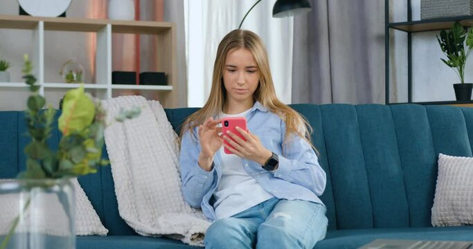 Portrait of young woman in casual clothes using her smartphone resting at home on comfortable sofa