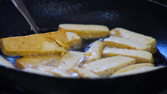 Turning frying tofu slices in hot oil with fork in fry pan on kitchen stove close up