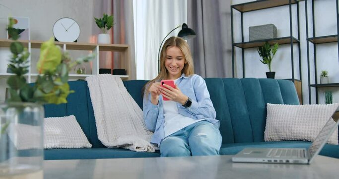 Smiling teen woman in casual clothes revision funny videos on phone sitting on the sofa in modern living-room