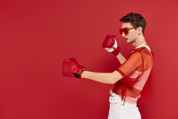 fashionable androgynous man with red fishnet posing actively with boxing gloves and looking away