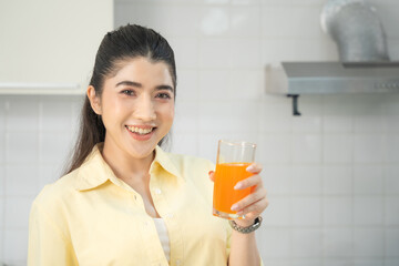 Smiling woman in a yellow shirt holds a glass of orange juice, standing in a well-lit kitchen, embodying a fresh start to the day.