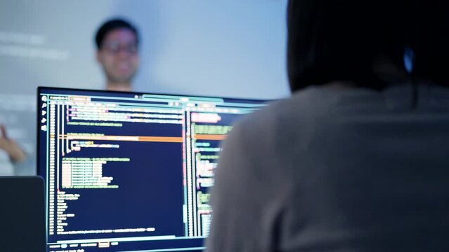 A male tech specialist gestures while explaining coding logic to a colleague against the backdrop of computer screens with code.