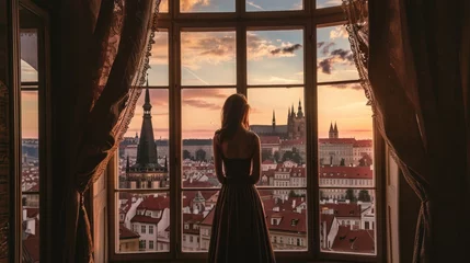 Poster A graceful lady standing by a large window with a view of historic buildings in the city of Prague, Czech Republic in Europe. © rabbit75_fot