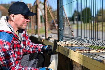 A man in overalls and gloves is repairing the fence in front of the family house. Close-up view and blurred background