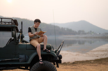 A young man contemplates sitting in his off-road vehicle by the lake. holding a notebook in the...