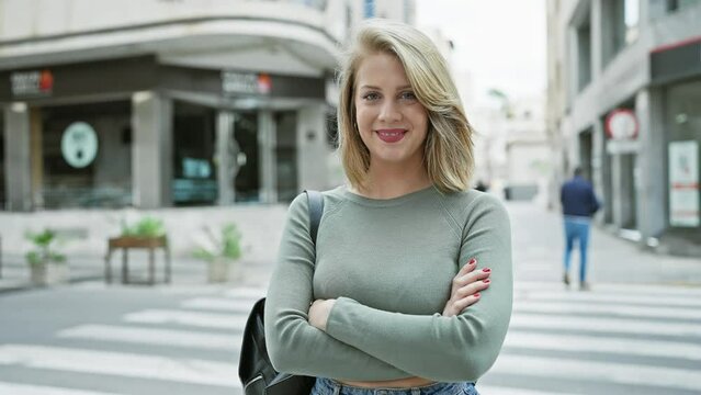 Smiling young woman with short blonde hair crossing arms while standing on a city street, portraying a casual urban lifestyle.