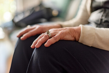 An elderly woman sits on a couch with her hands resting on her knees, wearing a ring on her left...