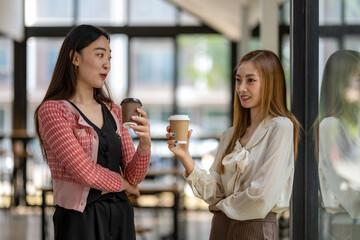Two female coworkers enjoy a casual conversation while holding coffee cups during a break in the office corridor.