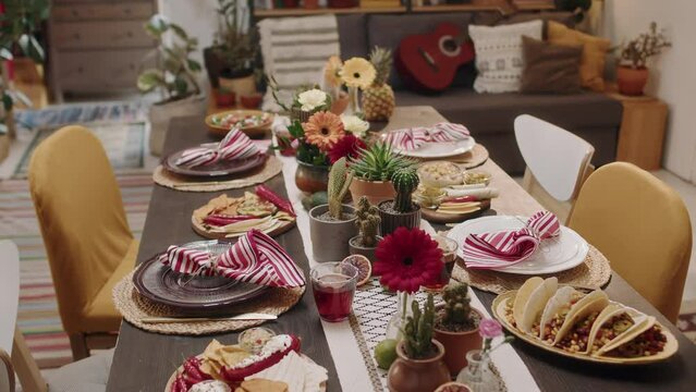 Close-up dynamic no people shot of dinner table with scrumptious traditional Mexican food and snacks, ready for party - nachos, tacos, chalupa, salsa, guacamole, salad, decorated with flowers, cacti