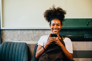 Portrait of a smiling black barista also the coffee shop owner using a mobile phone. Working in uniform communicating happily. Entrepreneur's relaxation is evident. - obrazy, fototapety, plakaty