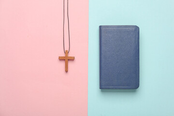 Bible book and wooden Christian cross on a string, pink blue background