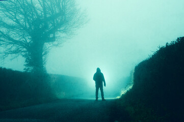 A spooky hooded figure. Standing on a haunted road. On a foggy winters night in the countryside.