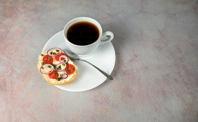 Coffee in white cup with bagel and cream cheese, tomatoes, capers and red onion on a white plate against a pink tinted counter cop