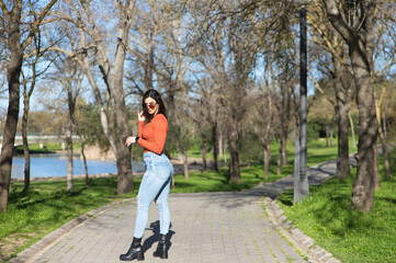 Young and beautiful brunette woman dressed in casual clothes and sunglasses, walks along a path between deciduous trees in autumn, in the background a lake.