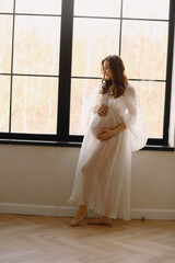 Portrait of beautiful pregnant woman in stylish white dress posing near windows in the studio