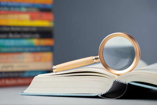 Magnifying glass on an opened book with colorful books pile in the background, searching information, glossary