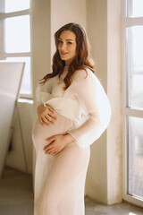 Portrait of brunette pregnant woman in stylish ivory dress in the studio. Stylish pregnancy concep